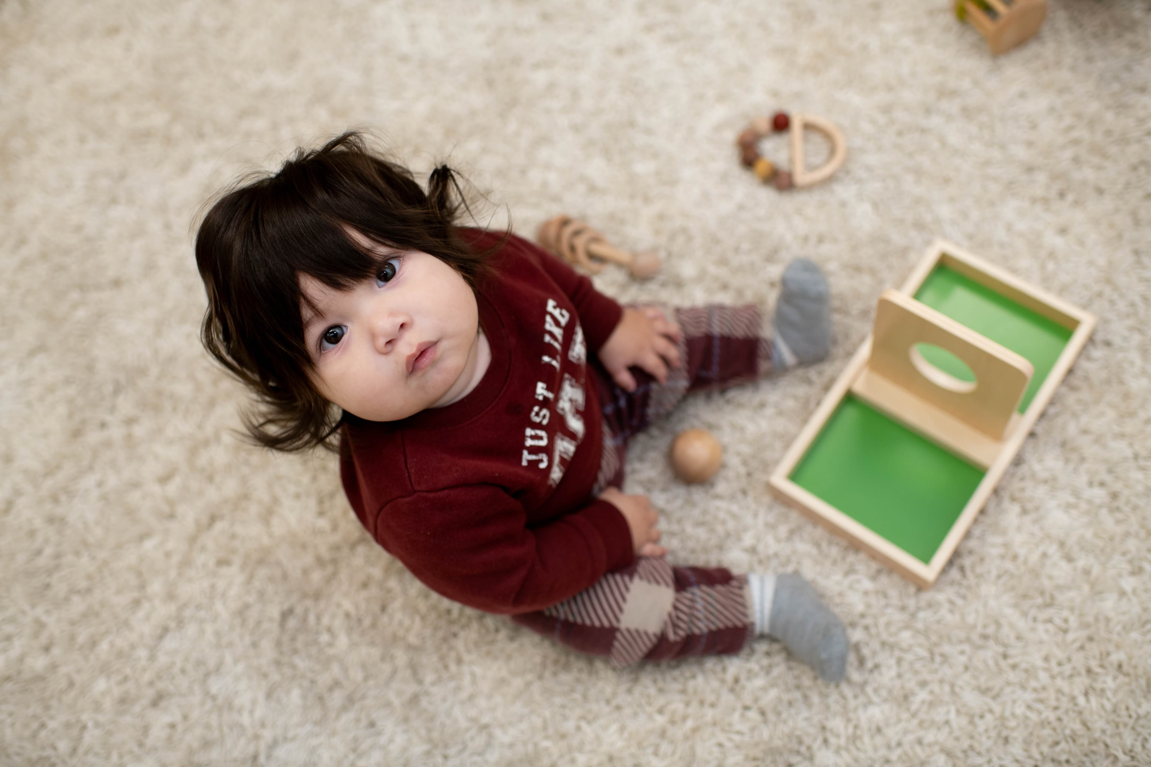 A baby sits on a soft rug beside a wooden box-and-ball material.